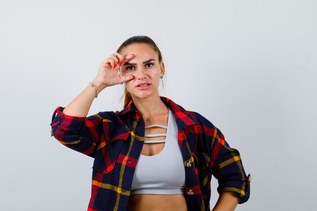 Junge Frau in bauchfreiem Top, kariertem Hemd, das durch die Finger schaut und hübsch aussieht, Vorderansicht.