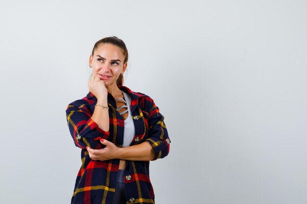 Junge Frau in bauchfreiem Top, kariertem Hemd, das in denkender Pose steht und nachdenklich aussieht, Vorderansicht.