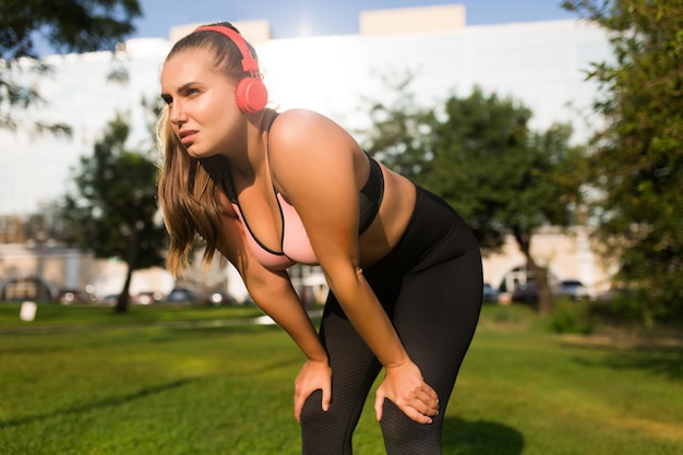 Junge Frau in Übergröße in sportlichem Oberteil und Leggings mit roten Kopfhörern, die müde zur Seite schaut, während sie sich im Stadtpark auf die Knie stützt