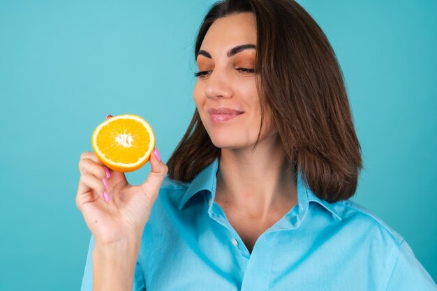 Junge Frau in blauem Hemd an der Wand hält eine Orange, posiert fröhlich, gut gelaunt, lacht, lächelt