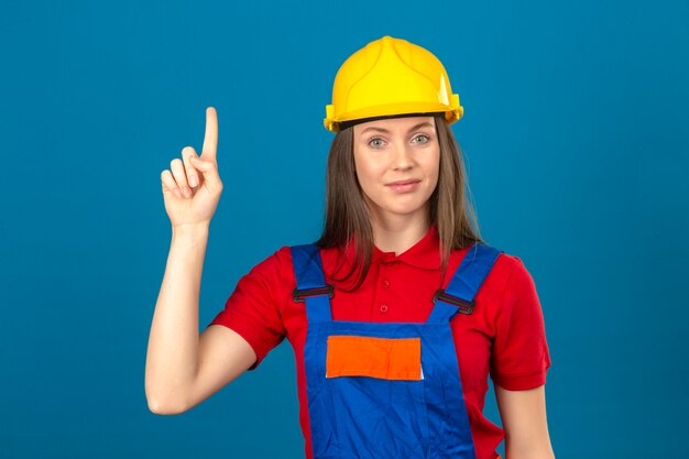 Junge Frau in der Bauuniform und im gelben Sicherheitshelm, der neue Idee zeigt, Finger oben auf blauem Hintergrund stehend