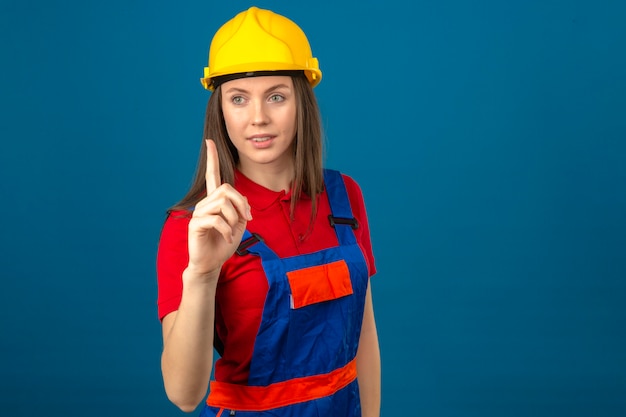 Junge Frau in der Bauuniform und im gelben Sicherheitshelm, der oben mit dem lächelnden Finger auf blauem Hintergrund zeigt