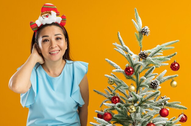 junge Frau in der blauen Spitze, die lustige Weihnachtsfelge auf dem Kopf betrachtet Kamera betrachtet, die mit glücklichem Gesicht steht, das neben einem Weihnachtsbaum über orange Hintergrund steht