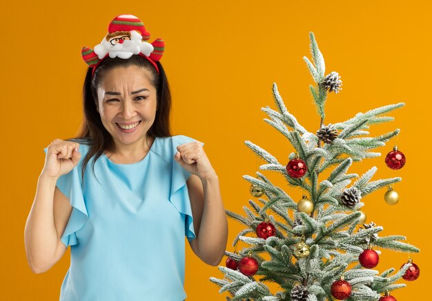 junge Frau in der blauen Spitze, die lustige Weihnachtsfelge auf dem Kopf trägt geballte Fäuste glücklich und aufgeregt steht neben einem Weihnachtsbaum über orange Hintergrund
