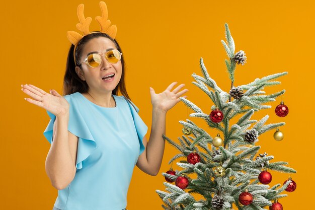 Junge Frau in der blauen Spitze, die lustigen Rand mit Hirschhörnern und gelben Gläsern trägt, die Kamera glücklich und aufgeregt stehen, die neben einem Weihnachtsbaum über orange Hintergrund stehen