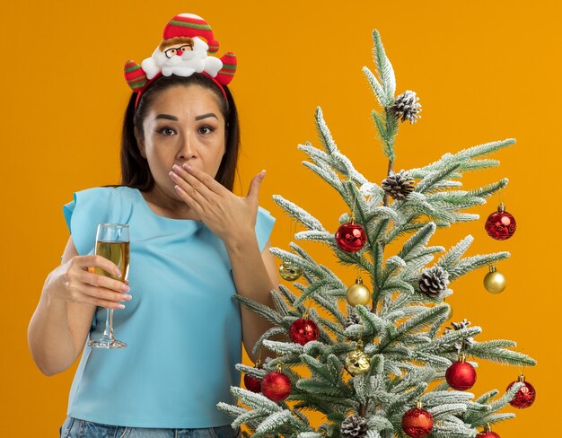 junge Frau in der blauen Spitze, die lustigen Weihnachtsrand auf Kopf hält, der Glas Champagner betrachtet, der Kamera betrachtet, die schockiert ist, den Mund mit der Hand stehend, die neben einem Weihnachtsbaum über orange Hintergrund steht