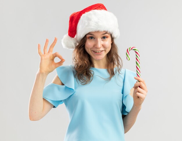 Junge Frau in der blauen Spitze und in der Weihnachtsmannmütze, die Zuckerstange hält, die glücklich und positiv schaut, ok Zeichen zeigend