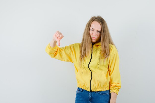 Junge Frau in der gelben Bomberjacke und in der blauen Jeans, die Daumen nach unten zeigen und unzufrieden aussehen, Vorderansicht.