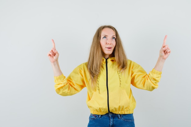 Junge Frau in der gelben Bomberjacke und in der blauen Jeans, die oben zeigen und glücklich schauen, Vorderansicht.