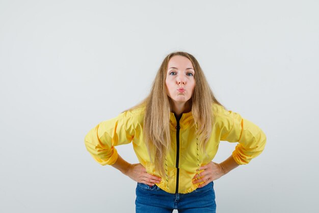 Junge Frau in der gelben Bomberjacke und in der blauen Jeans, die sich biegt, Hände auf Taille hält und Küsse zur Kamera sendet und optimistisch, Vorderansicht schaut.