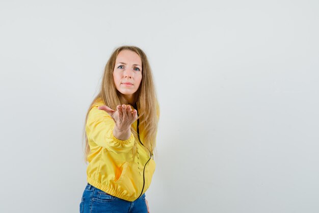 Junge Frau in der gelben Bomberjacke und in der blauen Jeans streckt Hand zur Kamera und schaut überrascht, Vorderansicht.