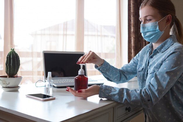 Junge Frau in der Gesichtsmaske, die Gadget-Oberflächen an ihrem Arbeitsplatz desinfiziert