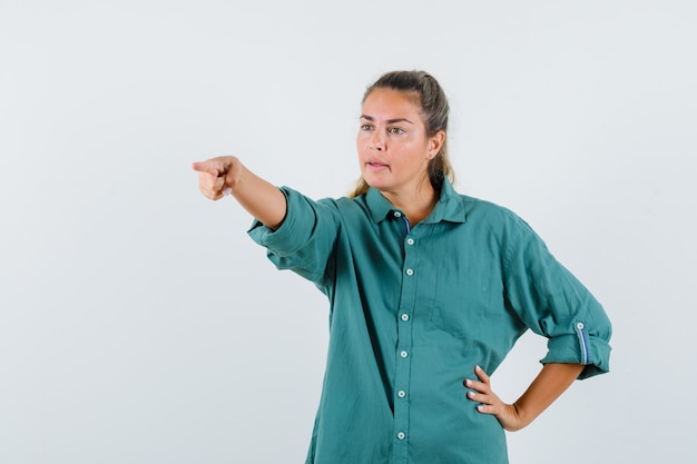 Junge Frau in der grünen Bluse, die mit Zeigefinger nach links zeigt, während sie eine andere Hand auf Taille hält und niedlich aussieht