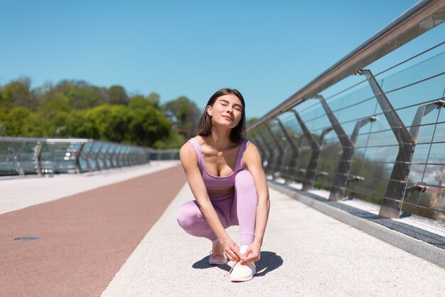 Junge Frau in der passenden Sportkleidung auf der Brücke am heißen sonnigen Morgen bindet Schnürsenkel