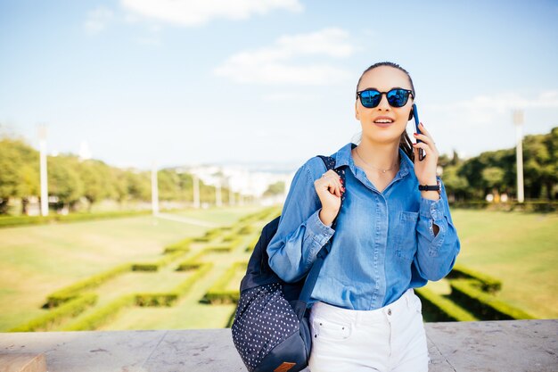 Junge Frau in der Sonnenbrille, die auf einem Handy gegen einen Pflanzenhintergrund spricht