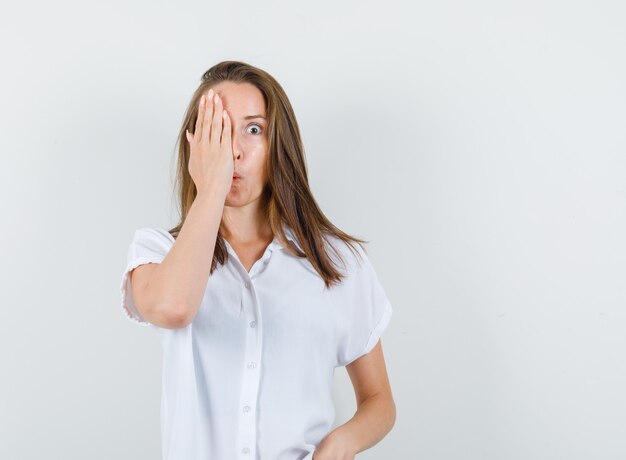 Junge Frau in der weißen Bluse, die sie auf Auge mit Hand schließt, während sie Lippen schmollt und verrückt aussieht