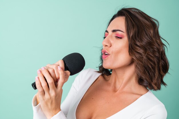 Junge Frau in einem weißen Abendkleid auf hellgrün mit leuchtend rosa Make-up hält ein Mikrofon, singt Karaoke