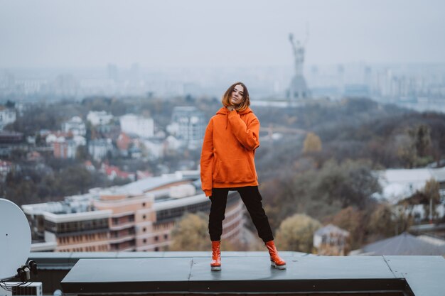 Junge Frau in einer orange Jacke posiert auf dem Dach eines Gebäudes im Stadtzentrum