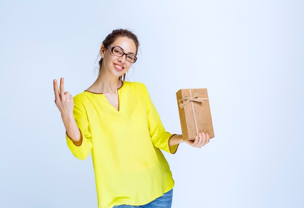 Junge Frau in gelbem Hemd, die eine Geschenkbox aus Karton hält und Freudenhandzeichen zeigt