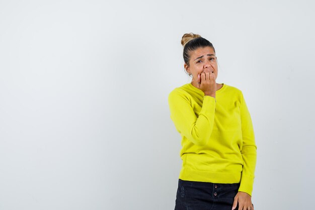 Junge Frau in gelbem Pullover und schwarzer Hose, die den Mund mit der Hand bedeckt, die Faust beißt und gehetzt aussieht looking