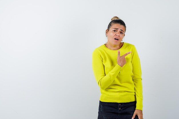 Junge Frau in gelbem Pullover und schwarzer Hose, die mit dem Zeigefinger nach rechts zeigt, eine Grimasse verzieht und aufgeregt aussieht