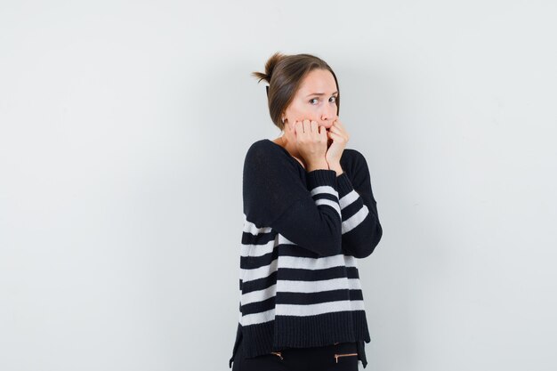 Junge Frau in gestreiften Strickwaren und schwarzen Hosen, die Fäuste beißen und erschrocken aussehen