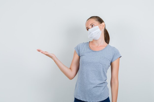 Junge Frau in grauem T-Shirt, Maske, Jeans, die mit Handflächenaufstrich beiseite schauen, Vorderansicht.