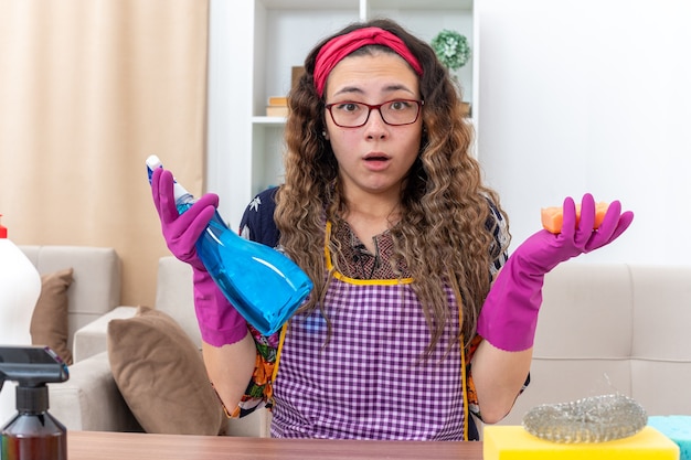 Junge Frau in Gummihandschuhen mit Reinigungsspray und Schwamm überrascht am Tisch sitzend mit Reinigungsmitteln und Werkzeugen im hellen Wohnzimmer