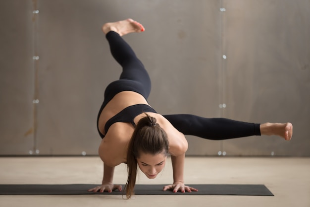 Junge Frau in Haltung Eka Pada Koundinyasana 1, graues Studio