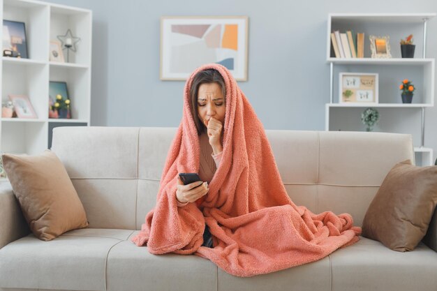 Junge Frau in Hauskleidung, die auf einer Couch unter einer Decke sitzt und eine SMS mit dem Smartphone schreibt, sieht unwohl aus und verbringt Zeit zu Hause