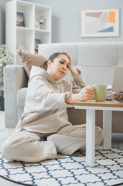 Junge Frau in Hauskleidung sitzt auf einem Boden in der Nähe der Couch zu Hause mit Laptop und einer Tasse Kaffee und hat ein Online-Training, das müde und gelangweilt aussieht