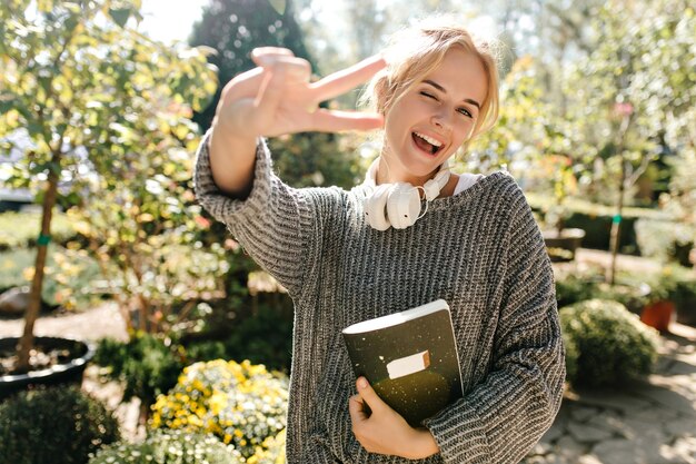 Junge Frau in Hochstimmung lächelt, zwinkert und zeigt ein Zeichen des Friedens Schnappschuss eines Mädchens mit Notizbuch, das im botanischen Garten posiert