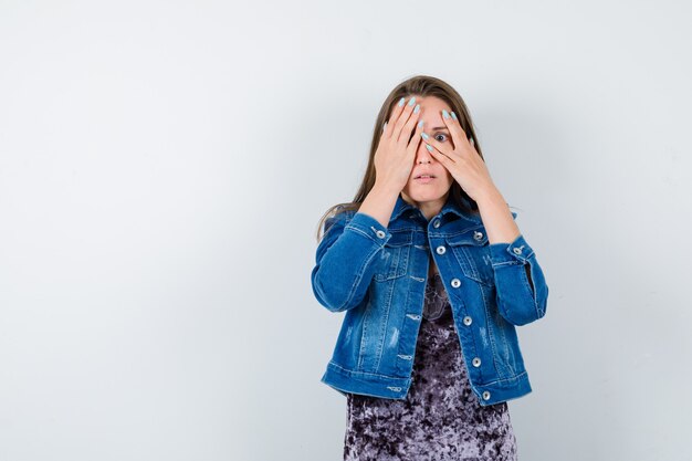 Junge Frau in Jeansjacke, die durch die Finger schaut und verängstigt aussieht, Vorderansicht.