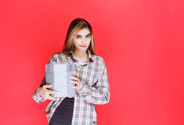 Junge Frau in kariertem Hemd, die eine silberne Geschenkbox hält und verwirrt und nachdenklich aussieht