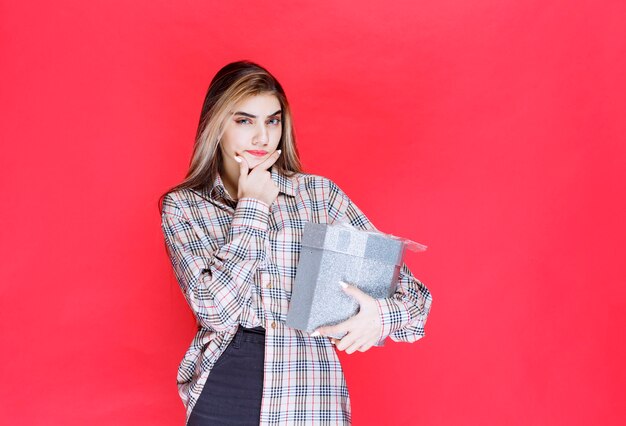 Junge Frau in kariertem Hemd, die eine silberne Geschenkbox hält und verwirrt und nachdenklich aussieht