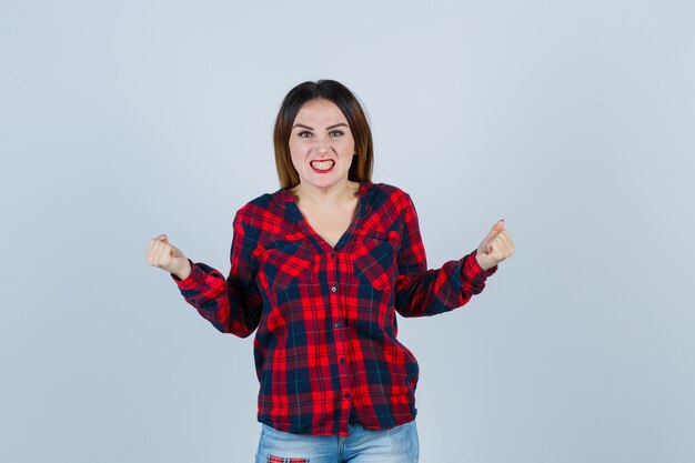 Junge Frau in kariertem Hemd, Jeans, die Fäuste ballt und wütend aussieht, Vorderansicht.