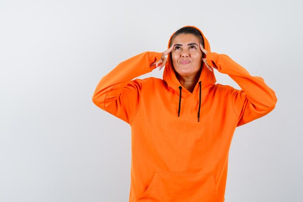 Junge Frau in orangefarbenem Hoodie, die Finger auf die Schläfen legt, nach oben schaut und über etwas nachdenkt und nachdenklich aussieht