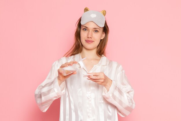 junge Frau in Pyjama und Schlafmaske mit Make-up-Spray und Lächeln auf rosa
