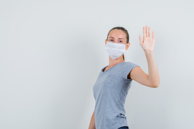 Junge Frau in T-Shirt, Maske, Jeans, die Hand winkt, um sich zu verabschieden und fröhlich auszusehen.