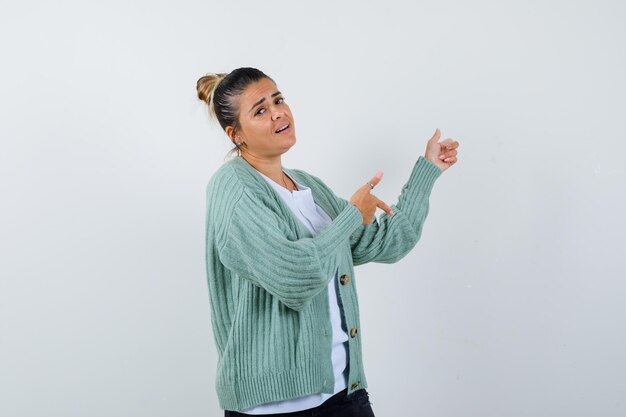 Junge Frau in weißem T-Shirt und mintgrüner Strickjacke, die mit den Zeigefingern nach rechts zeigt und genervt aussieht