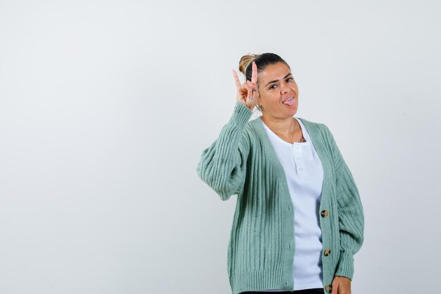 Junge Frau in weißem T-Shirt und mintgrüner Strickjacke, die Rock'n'Roll-Geste zeigt, die Zunge herausstreckt und glücklich aussieht