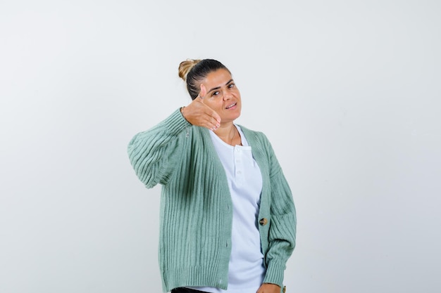 Junge Frau in weißem T-Shirt und mintgrüner Strickjacke, die zum Kommen einlädt und glücklich aussieht looking