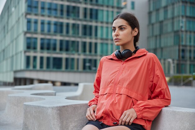 Junge Frau in Windjacke lehnt sich mit Ellbogen auf Stein und macht Übungen im Freien, motiviert zu einem gesunden Lebensstil und treibt Sport auf der Stadtstraße schaut nach vorne posiert gegen das städtische Gebäude dahinter