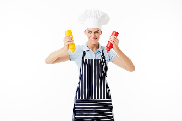 Junge Frau kocht in gestreifter Schürze und weißem Hut und blickt spielerisch in die Kamera, während sie Flaschen mit Ketchup und Senf in den Händen vor weißem Hintergrund hält