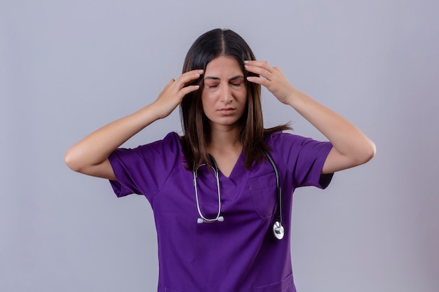 Junge Frau Krankenschwester, die Uniform trägt und mit Stethoskop müde aussehende Schläfen mit starken Kopfschmerzen sucht
