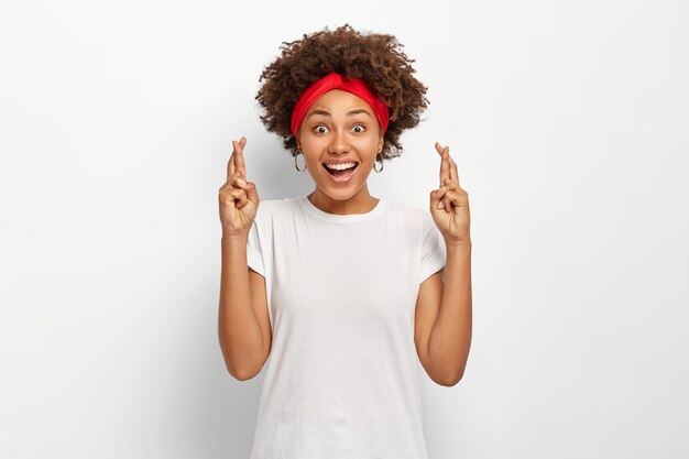 Junge Frau mit Afro-Frisur, drückt die Daumen für viel Glück, wartet auf Wunder, trägt rotes Stirnband und lässiges T-Shirt