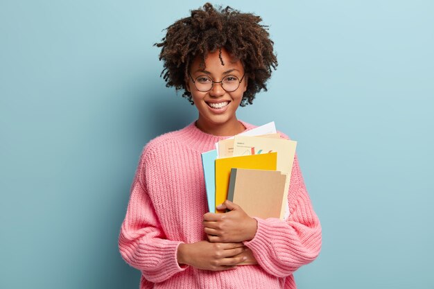 Junge Frau mit Afro-Haarschnitt, der rosa Pullover trägt und Lehrbücher hält