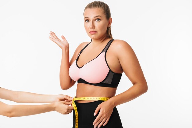 Junge Frau mit Übergewicht im sportlichen Oberteil, die erstaunt in die Kamera schaut, während sie die Taille auf weißem Hintergrund misst