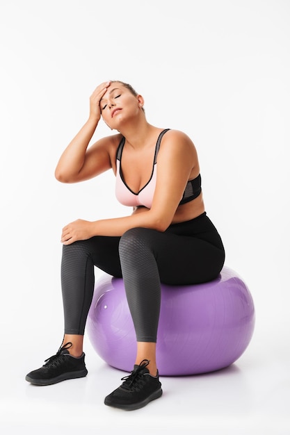 Junge Frau mit Übergewicht in sportlichem Oberteil und Leggings sitzt auf Fitnessball, während sie müde die Augen vor weißem Hintergrund schließt