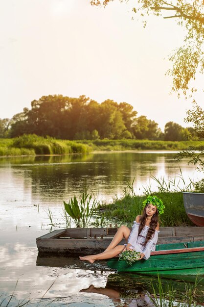 Junge Frau mit Blumenkranz auf dem Kopf, entspannt auf dem Boot auf dem Fluss bei Sonnenuntergang. Schöner Körper und Gesicht. Fantasy-Kunstfotografie. Konzept der weiblichen Schönheit, Ruhe im Dorf